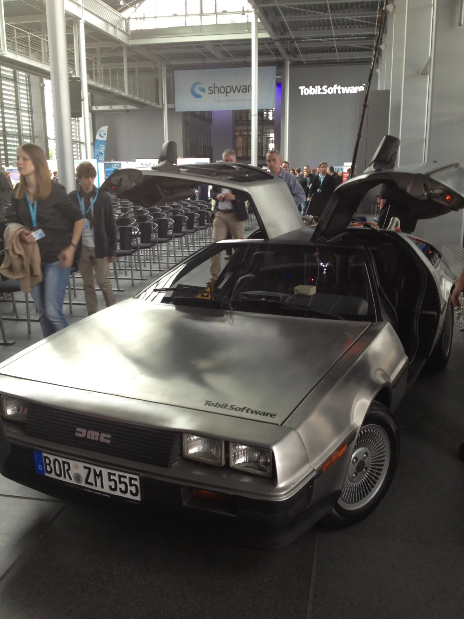 Der DeLorean von vorn mit beidseitig geöffneten Flügeltüren, auf der Haube der Schriftzug Tobit.Software, im Hintergrund ein Shopware-Banner