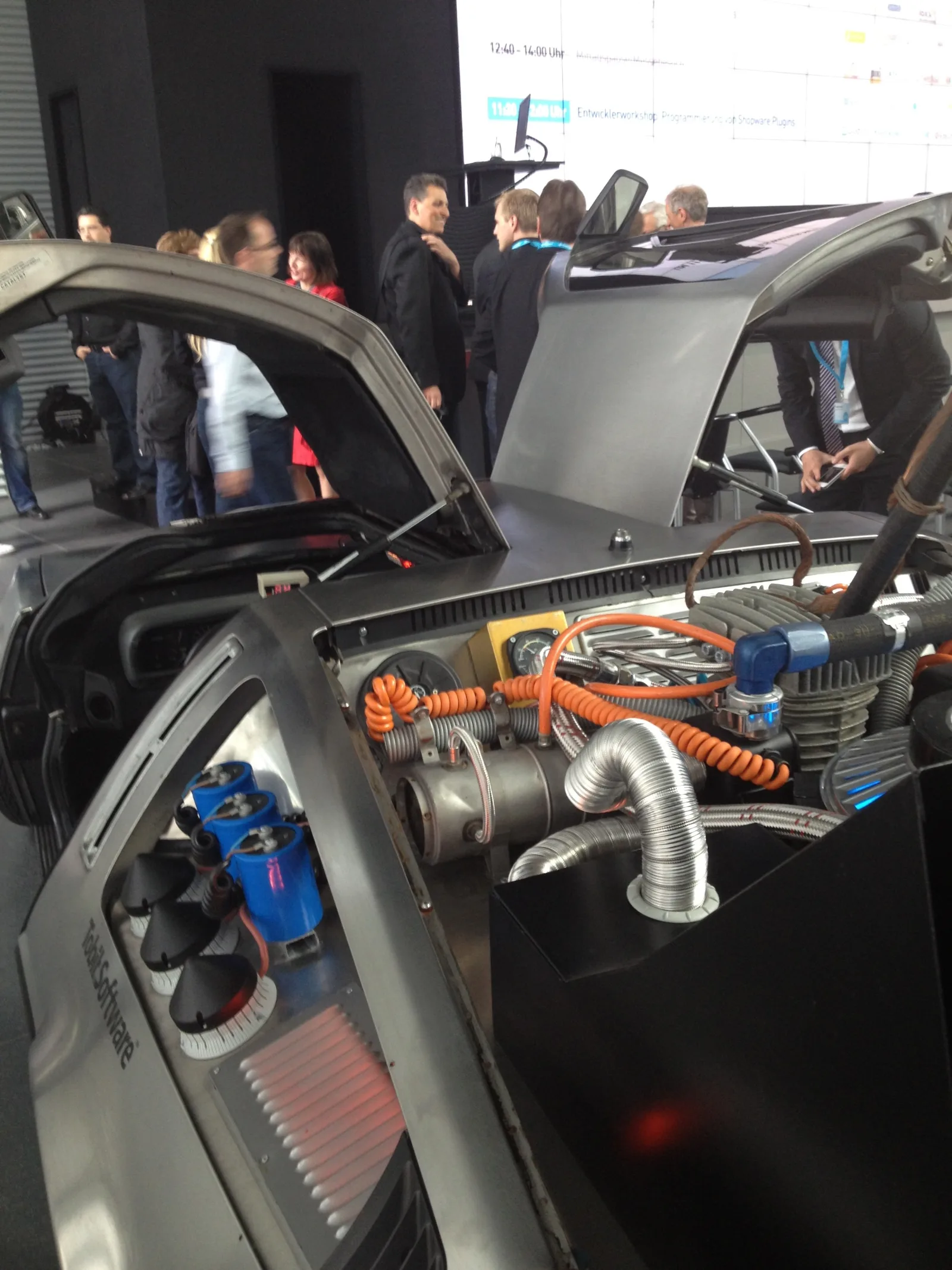 Geöffneter Heckbereich des DeLorean mit sichtbarer Technik aus Kabeln, Kondensatoren und Schläuchen, im Hintergrund die Programmtafel mit dem Eintrag Entwicklerworkshop Programmierung von Shopware Plugins