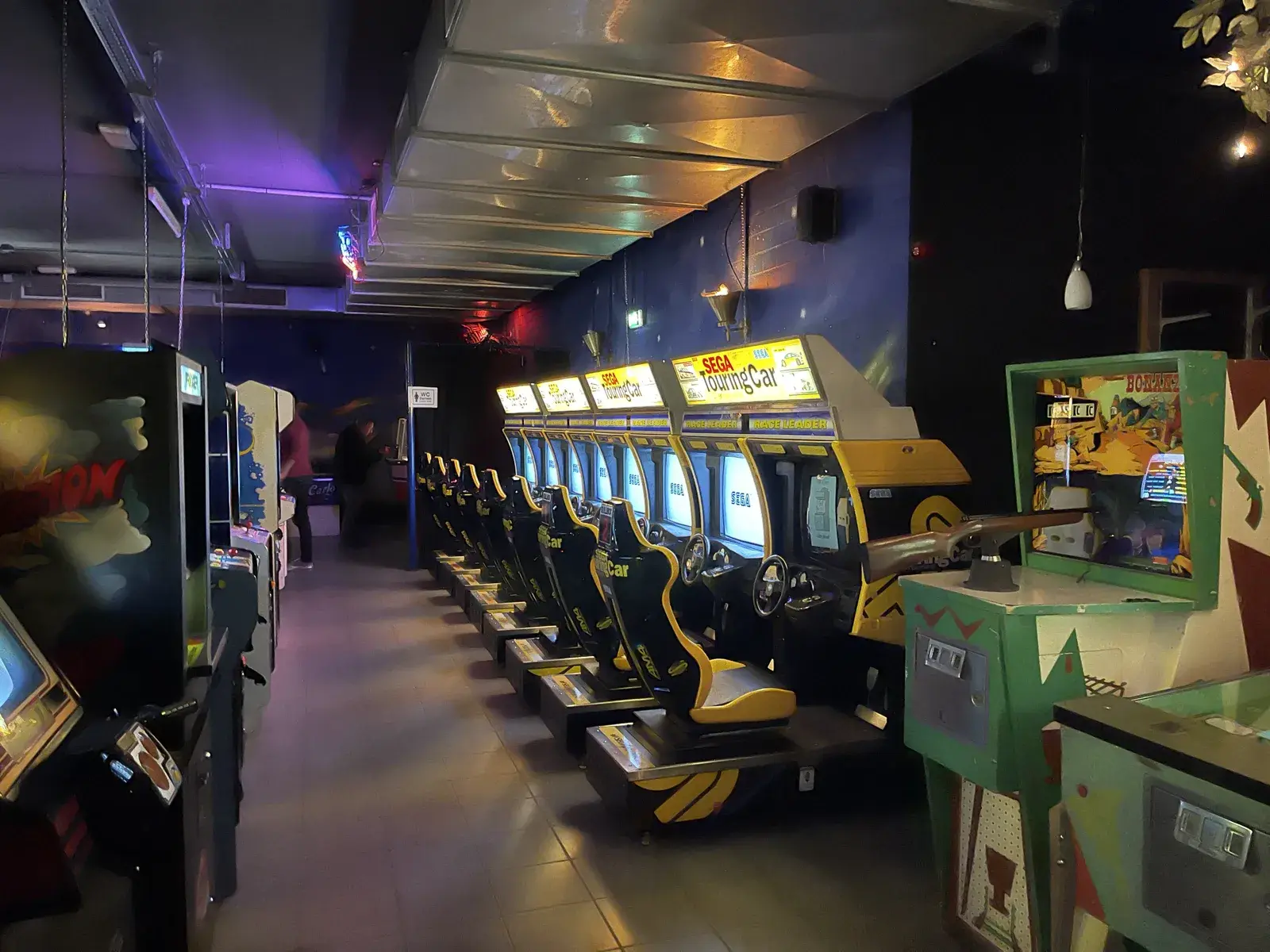 Reihe von SEGA-Touring-Car-Automaten mit Sitzen und Lenkrädern, rechts ein Schießautomat