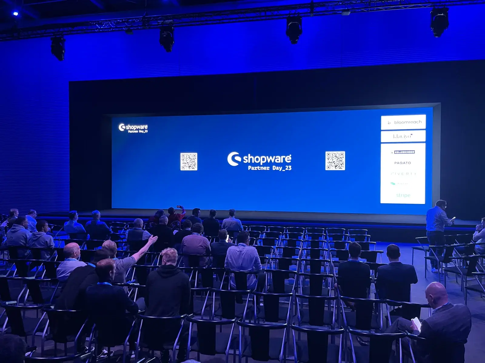 Bühne des Shopware Partner Day 2023 mit blauer Leinwand, Sponsorenlogos und Publikum im Saal